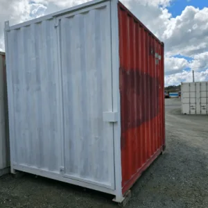Used 10ft container part painted grey and part red, standing on gravel under a cloudy sky