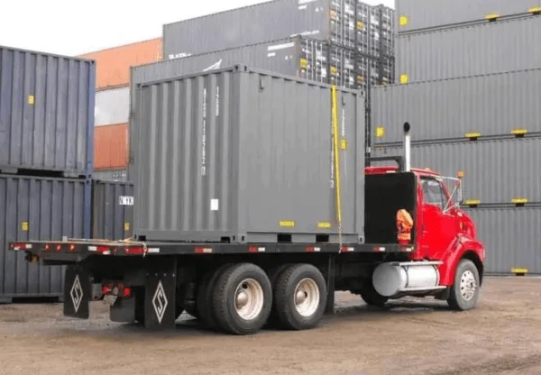 Grey 10ft container loaded on the bed of a red flatbed delivery truck at a container depot