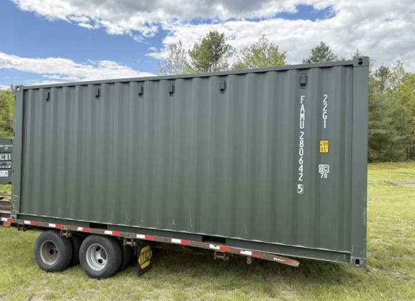 Green 20ft container loaded lengthways on a flatbed trailer parked on grass